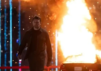 Homem de casaco escuro caminha à noite. Um carro em chamas está à direita, luzes azuis verticais à esquerda. Clima tenso de ação.