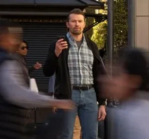 Homem barbudo sério de camisa xadrez e jaqueta em rua movimentada. Ele se destaca entre pessoas borradas, com olhar fixo.