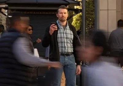 Homem barbudo sério de camisa xadrez e jaqueta em rua movimentada. Ele se destaca entre pessoas borradas, com olhar fixo.