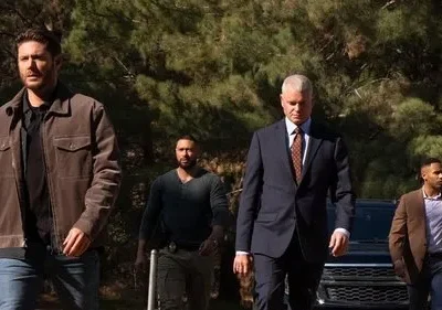 Quatro homens sérios, de várias etnias, caminham com determinação. Um de jaqueta marrom, outro de terno. Floresta e SUV ao fundo.