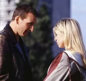 Um homem de jaqueta sorri levemente para uma mulher loira séria. Encontro urbano diurno com clima intrigante.