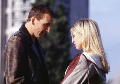 Um homem de jaqueta sorri levemente para uma mulher loira séria. Encontro urbano diurno com clima intrigante.