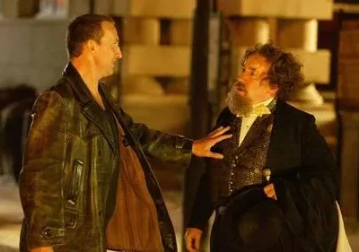 Dois homens em trajes de época em uma rua escura. Um, de jaqueta, toca o peito do outro, barbudo, que parece surpreso.