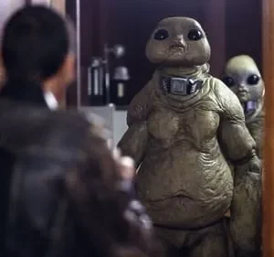 Um homem de costas olha para dois alienígenas com olhos grandes, um robusto e um menor atrás, em um ambiente interno escuro.