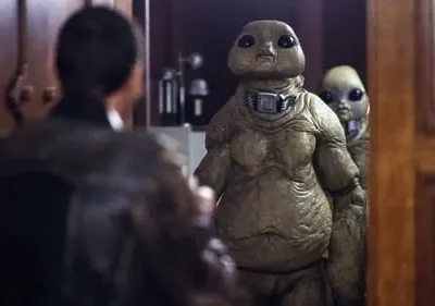 Um homem de costas olha para dois alienígenas com olhos grandes, um robusto e um menor atrás, em um ambiente interno escuro.