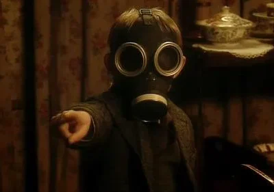 Criança com máscara de gás aponta para frente em sala escura e antiga. Ambiente misterioso e perturbador.