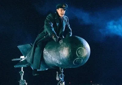 Homem em uniforme militar escuro cavalga euforicamente uma bomba com barbatanas, suspensa em um fundo escuro e nublado.