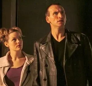 Homem de jaqueta de couro preta e mulher com coques no cabelo olham fixamente para cima em um ambiente escuro.