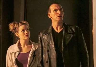 Homem de jaqueta de couro preta e mulher com coques no cabelo olham fixamente para cima em um ambiente escuro.