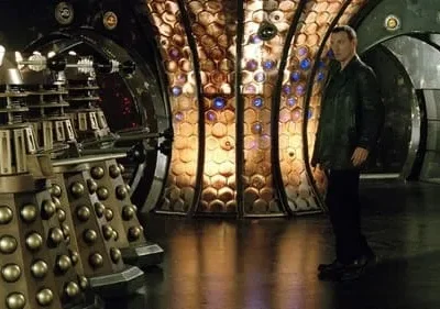 Robôs dourados de aparência ameaçadora em primeiro plano. Um homem de jaqueta escura olha. Cenário futurista com paredes iluminadas.