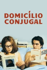 Mulher e homem jovens na cama,cobertos por um lençol azul,lendo livros. Ela usa óculos e ele a observa de soslaio.