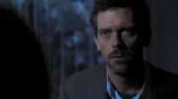 Homem de barba e cabelo escuro, olhos azuis intensos, blazer e camisa azul, olha fixamente em ambiente escuro. Clima sério/intenso.