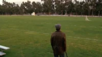 Um homem de boina e casaco, de costas, num vasto campo verde com um gol e árvores ao fundo. Cena calma e contemplativa.