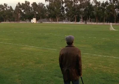 Um homem de boina e casaco, de costas, num vasto campo verde com um gol e árvores ao fundo. Cena calma e contemplativa.