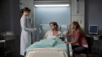 Médica em jaleco fala com paciente deitado em cama de hospital. Uma mulher sentada ao lado segura a mão do paciente. Cena dramática.