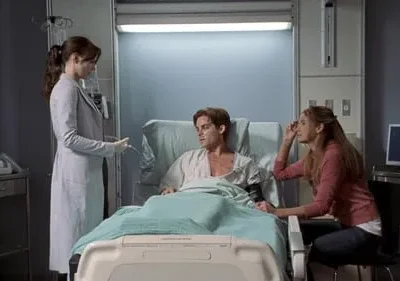 Médica em jaleco fala com paciente deitado em cama de hospital. Uma mulher sentada ao lado segura a mão do paciente. Cena dramática.