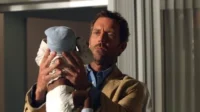 Um homem de barba e casaco claro segura um bebê de touca azul em um ambiente interno. Seu olhar é intenso e sério.