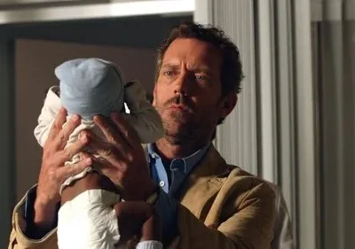 Um homem de barba e casaco claro segura um bebê de touca azul em um ambiente interno. Seu olhar é intenso e sério.