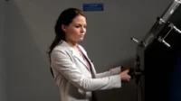 Mulher de cabelo escuro, com blazer claro e expressão séria, opera um aparelho científico ou máquina com tubos e botões em laboratório.