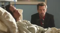 Mulher deitada em cama de hospital. Um homem sério com barba e bengala a observa ao fundo. Clima sombrio.