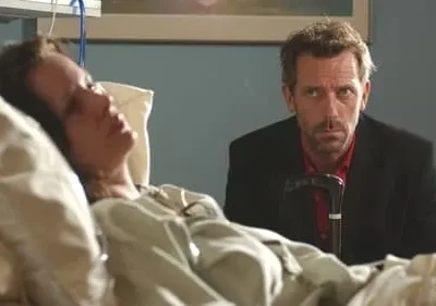 Mulher deitada em cama de hospital. Um homem sério com barba e bengala a observa ao fundo. Clima sombrio.