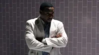 Homem negro em jaleco branco, braços cruzados, olhar sério. Fundo escuro de azulejos cinza, com luz lateral.
