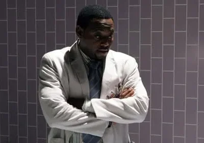 Homem negro em jaleco branco, braços cruzados, olhar sério. Fundo escuro de azulejos cinza, com luz lateral.