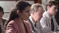 Mulher de blazer rosa e pérolas, atenta, ao lado de homens em jalecos brancos. Um clima profissional e sério.