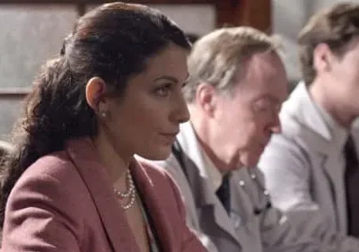 Mulher de blazer rosa e pérolas, atenta, ao lado de homens em jalecos brancos. Um clima profissional e sério.