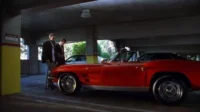 Dois homens sérios em um estacionamento. Um com bengala, outro de terno, ao lado de um carro esportivo vermelho vintage. Clima sombrio.