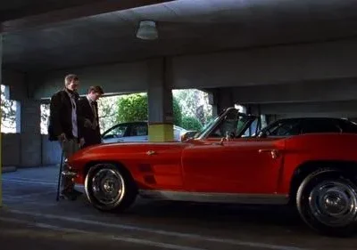 Dois homens sérios em um estacionamento. Um com bengala, outro de terno, ao lado de um carro esportivo vermelho vintage. Clima sombrio.