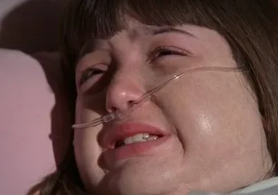 Mulher pálida em close-up, chorando intensamente com cânula nasal. Expressão de dor e angústia em leito.