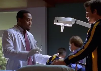 Médico sério de jaleco branco com seringa, ao lado de homem de casaco esportivo azul e amarelo, assistindo paciente em maca.