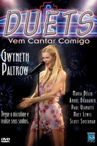 Mulher loira canta em microfone,vestido florido e brilhante,em palco escuro com luzes de fogos. Clima musical e festivo.