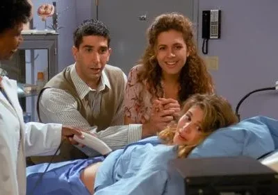 Médica faz ultrassom em mulher deitada na maca. Um homem observa preocupado, enquanto outra mulher sorri. Cenário de clínica médica com expectativa.