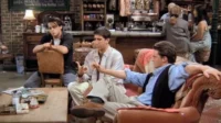 Três homens em um café aconchegante. Um gesticula, um reclinado no sofá e um sentado de costas na cadeira. Atmosfera descontraída.
