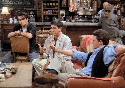 Três homens em um café aconchegante. Um gesticula, um reclinado no sofá e um sentado de costas na cadeira. Atmosfera descontraída.