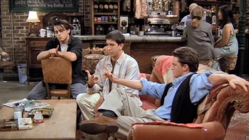 Três homens em um café aconchegante. Um gesticula,um reclinado no sofá e um sentado de costas na cadeira. Atmosfera descontraída.