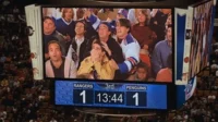 Jumbotron de estádio exibe grupo de amigos com expressões de choque e euforia. Placar 1-1 em jogo de hóquei cria suspense.