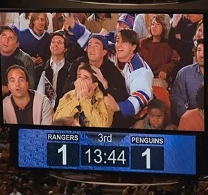 Jumbotron de estádio exibe grupo de amigos com expressões de choque e euforia. Placar 1-1 em jogo de hóquei cria suspense.