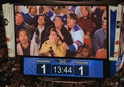 Jumbotron de estádio exibe grupo de amigos com expressões de choque e euforia. Placar 1-1 em jogo de hóquei cria suspense.