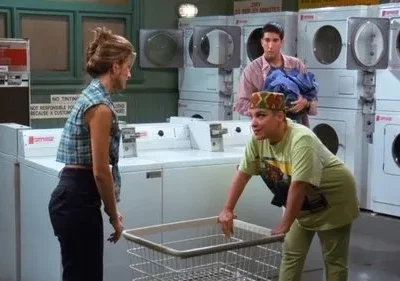 Três pessoas em uma lavanderia. Uma mulher com chapéu colorido se inclina sobre um cesto de roupa. Outra mulher e um homem com roupas a observam.