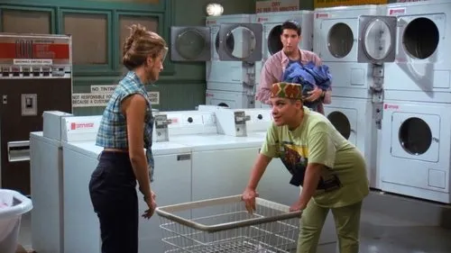 Três pessoas em uma lavanderia. Uma mulher com chapéu colorido se inclina sobre um cesto de roupa. Outra mulher e um homem com roupas a observam.
