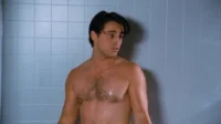 Homem molhado, sem camisa, cabelos escuros e expressão surpresa, em chuveiro de azulejos azuis.