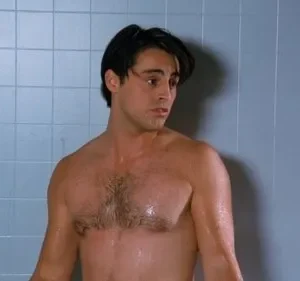 Homem molhado, sem camisa, cabelos escuros e expressão surpresa, em chuveiro de azulejos azuis.