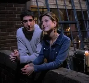 Um homem e uma mulher encostados em um parapeito, sob a luz de velas, com uma janela iluminada ao fundo. Clima íntimo e noturno.