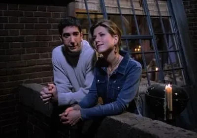 Um homem e uma mulher encostados em um parapeito, sob a luz de velas, com uma janela iluminada ao fundo. Clima íntimo e noturno.