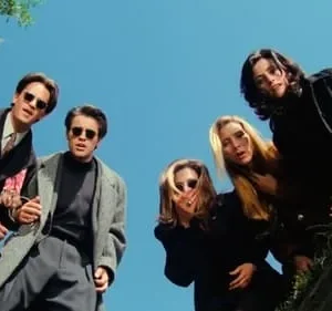 Seis amigos em casacos escuros e óculos escuros olham para baixo do alto de um buraco contra um céu azul.