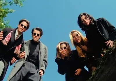 Seis amigos em casacos escuros e óculos escuros olham para baixo do alto de um buraco contra um céu azul.