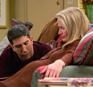 Homem moreno deitado em sofá verde conversa com mulher loira sentada, que olha para longe. Cenário de sala aconchegante.